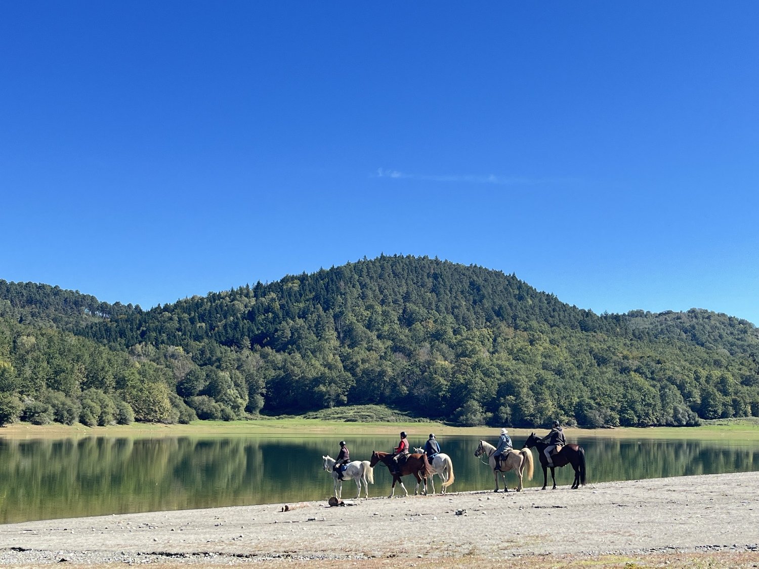 Wooded hills reflected in Lac de Mondely, with riders beside the water